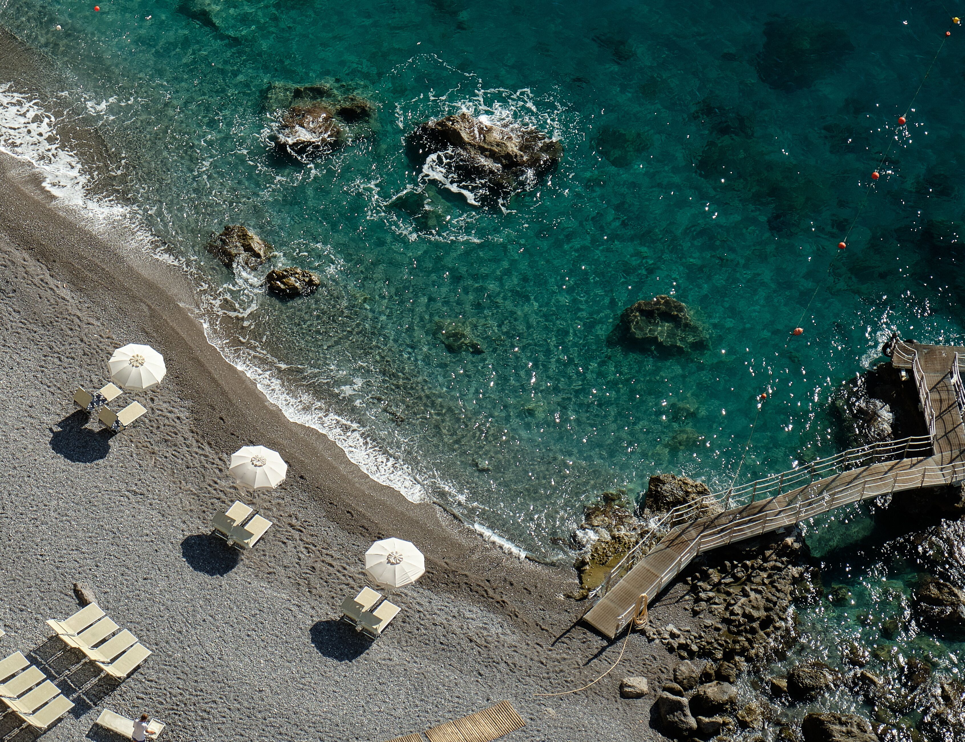Una playa con agua turquesa cristalina, afloramientos rocosos y un puente de madera.