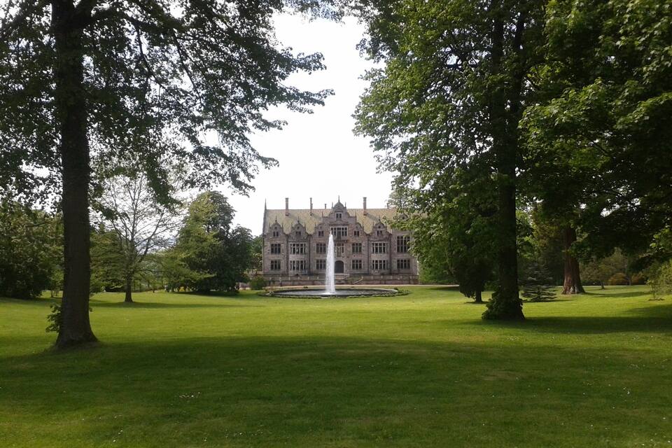 A large, historic building with a fountain in the center of a well-manicured lawn, surrounded by tall trees.