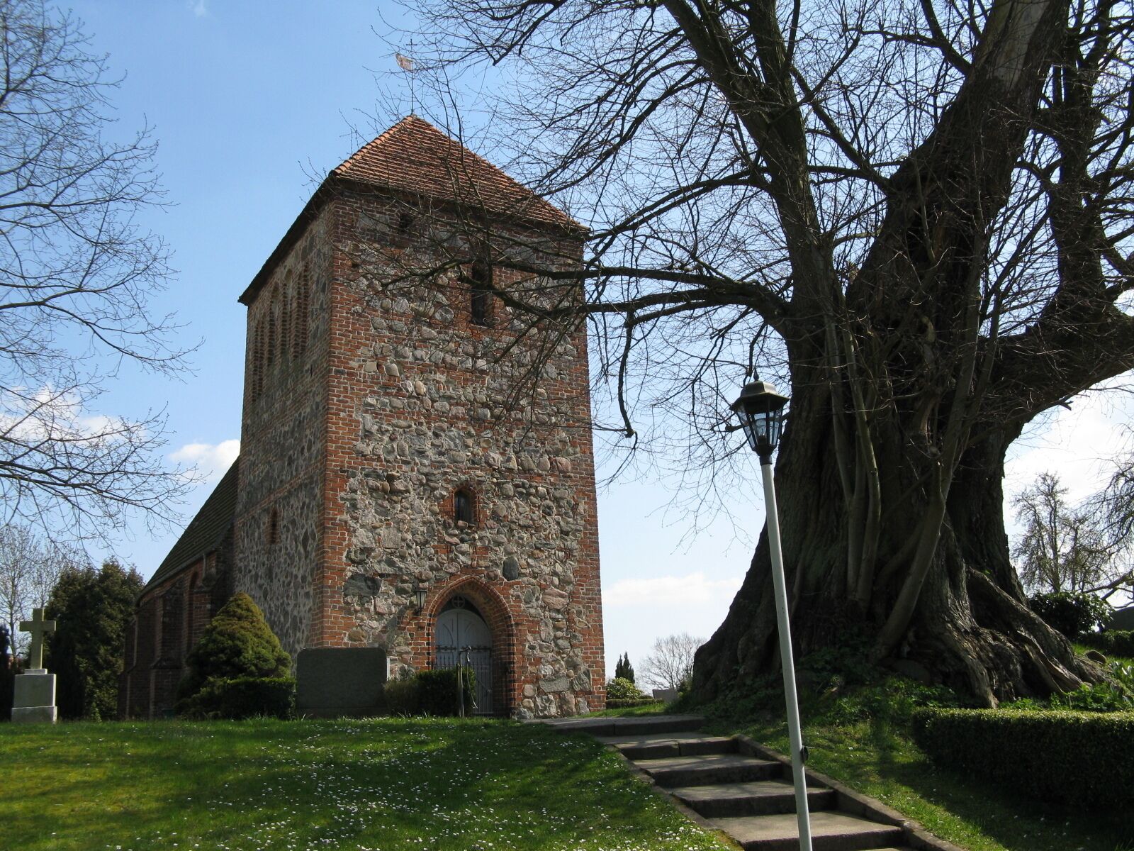 A stone church with a tiled roof, a large tree, and a pathway leading to the entrance.