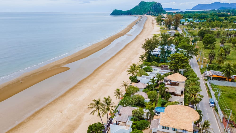 Une plage cĂŽtiĂšre offrant une vue dĂ©gagĂ©e sur lâocĂ©an, une rive sablonneuse et une zone rĂ©sidentielle avec des maisons et des palmiers.
