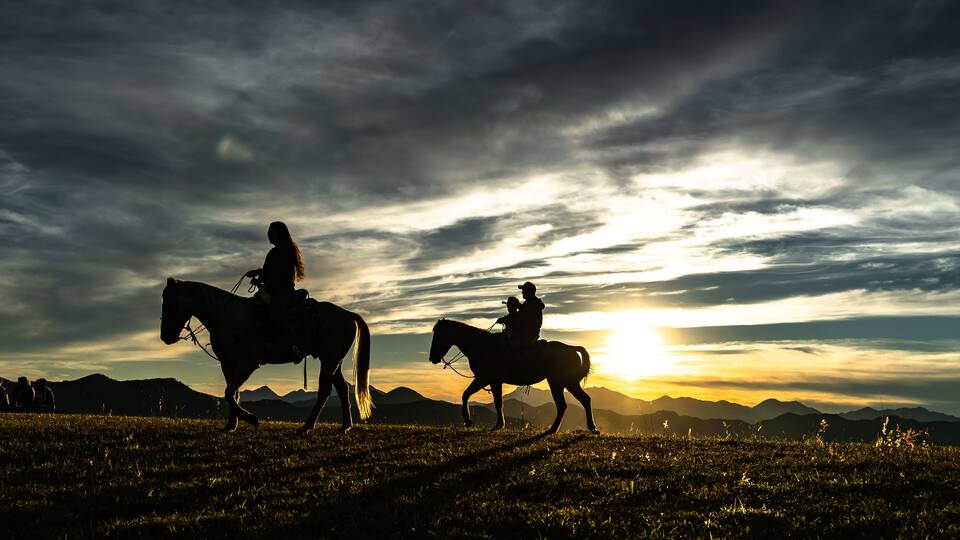 Two people riding horses on a grassy field with mountains in the background during sunset.