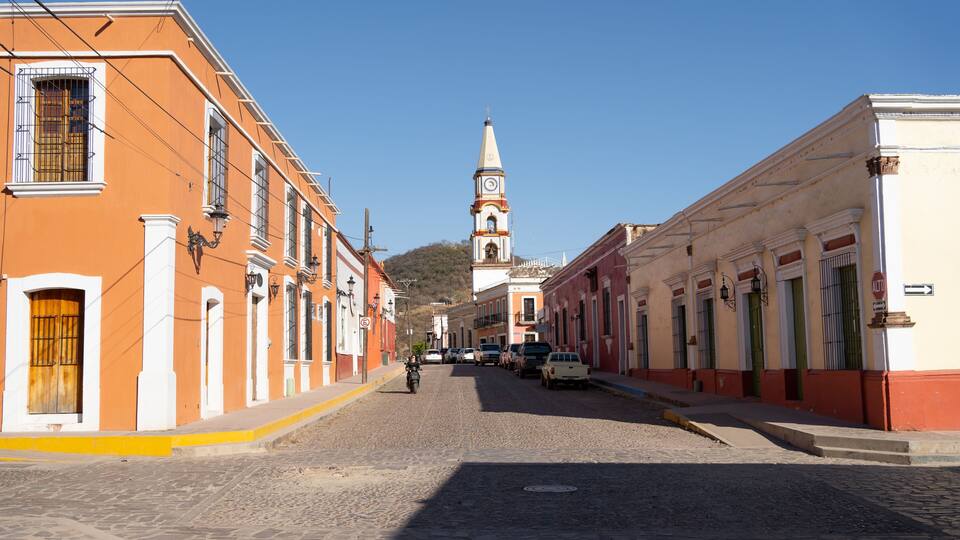 A cobblestone street lined with historic buildings, a church with a clock tower, and a clear blue sky.