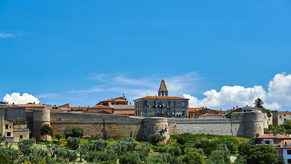 A historic stone wall with a tower, olive trees, and buildings with red-tiled roofs.