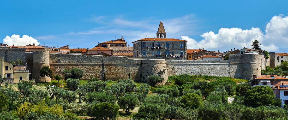 A historic stone wall with a tower, olive trees, and buildings with red-tiled roofs.