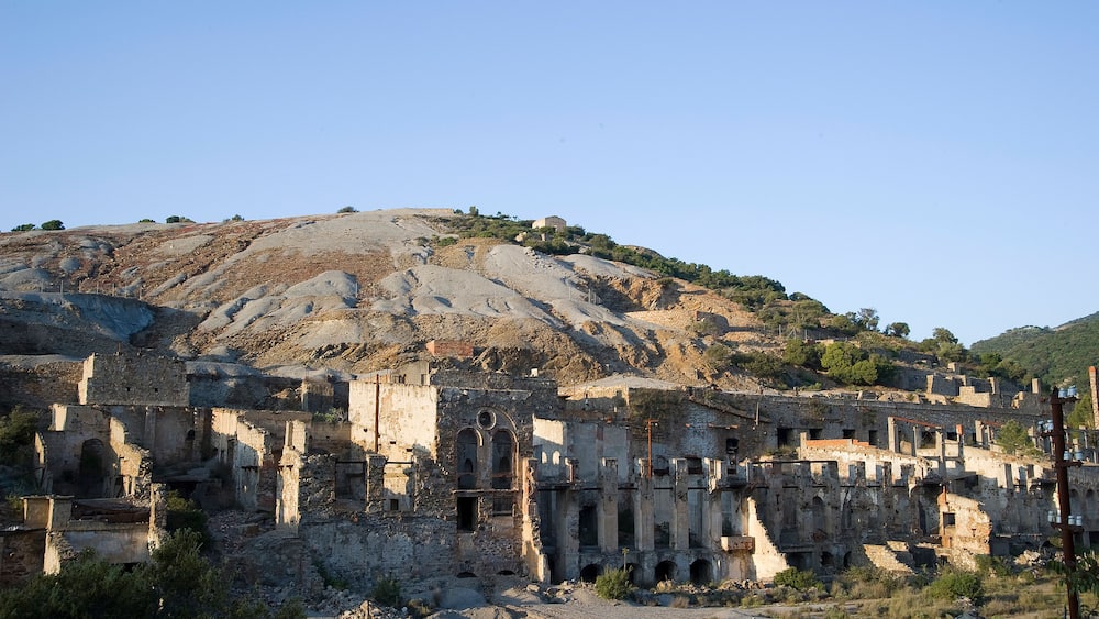 Des ruines antiques, avec des colonnes et des arches, une colline parsemée d’une végétation clairsemée et un ciel bleu dégagé.