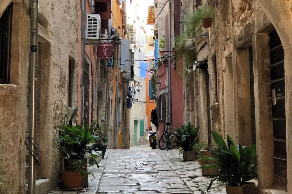 Uma rua de paralelepípedos estreita, ladeada por edifícios antigos, vasos com plantas e bicicletas.