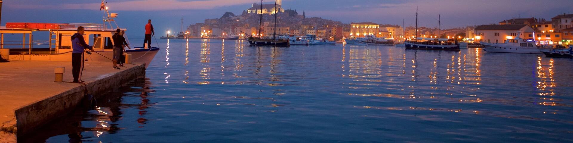 A waterfront at dusk with boats docked, a prominent tower, and buildings along the shore.