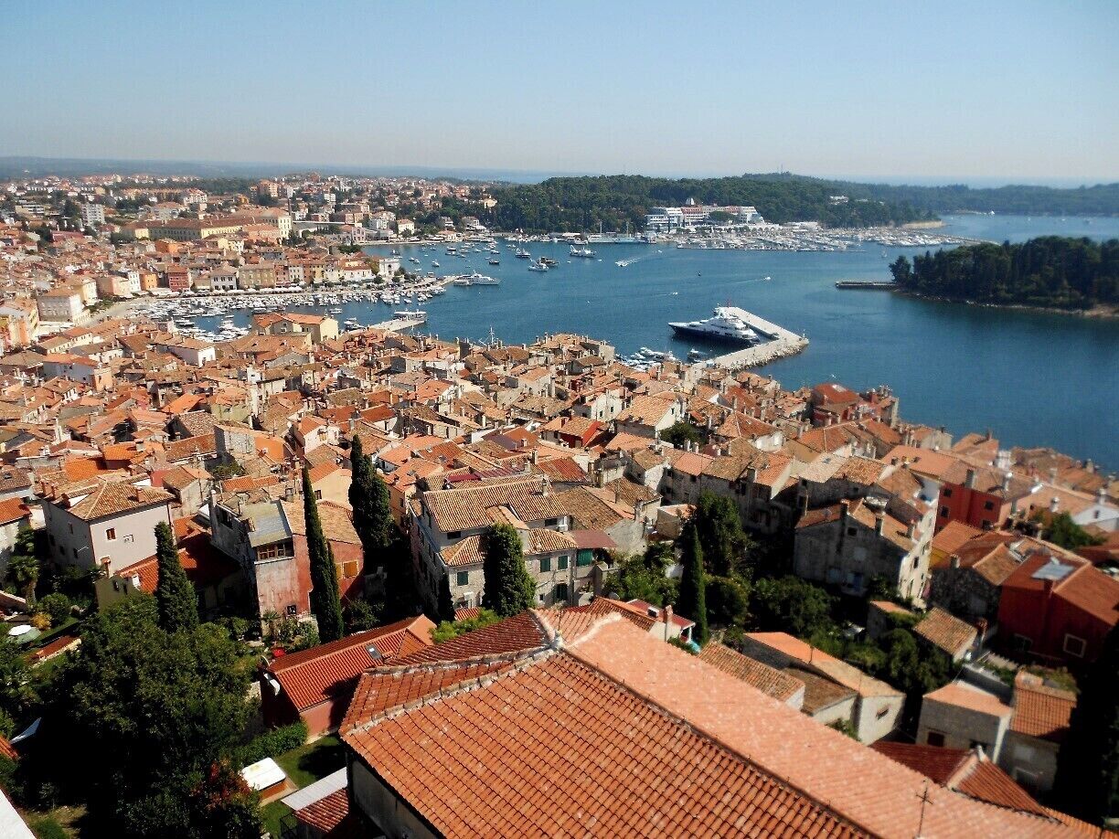 Une ville côtière aux bâtiments aux toits de tuiles rouges, un port de plaisance avec des bateaux et une mer d’un bleu limpide.