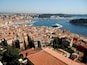 Un pueblo costero con edificios de tejados terracota, un puerto deportivo con barcos y un mar azul cristalino.