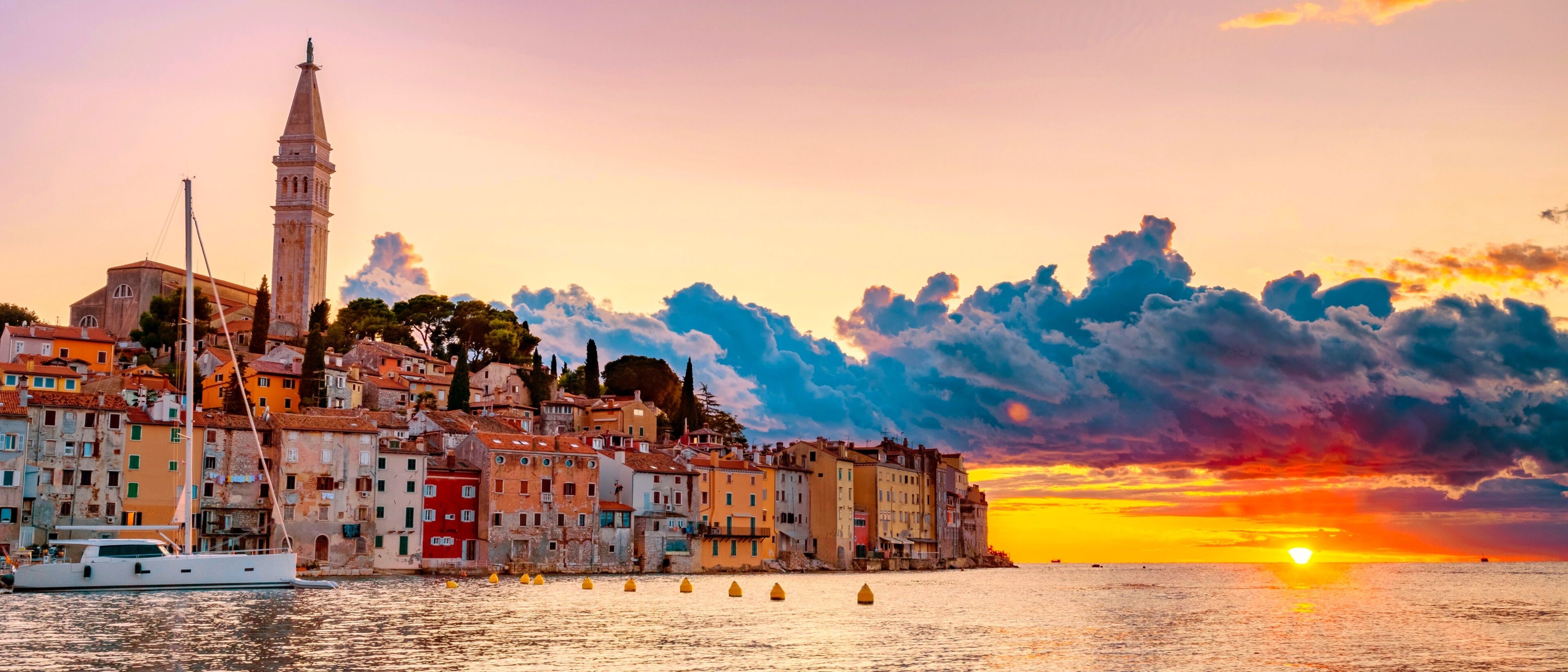 Un paisaje urbano costero al atardecer con una torre destacada, edificios coloridos y un mar tranquilo.