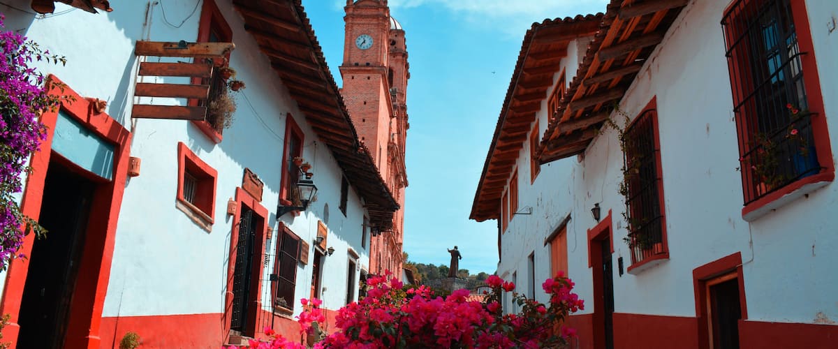 A narrow street flanked by white buildings with red accents, a prominent clock tower, and vibrant pink bougainvillea.