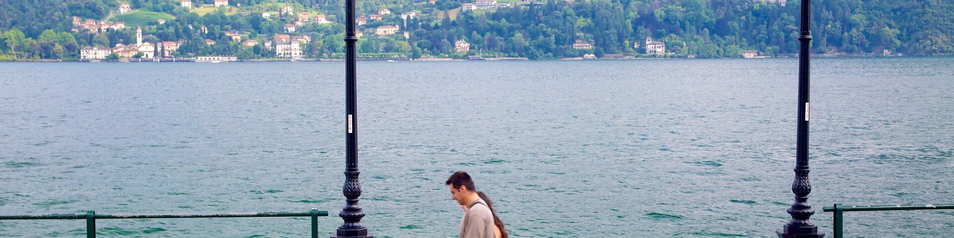 A waterfront promenade with a man walking, a woman in a pink dress, and a man in a blue shirt sitting on a bench.