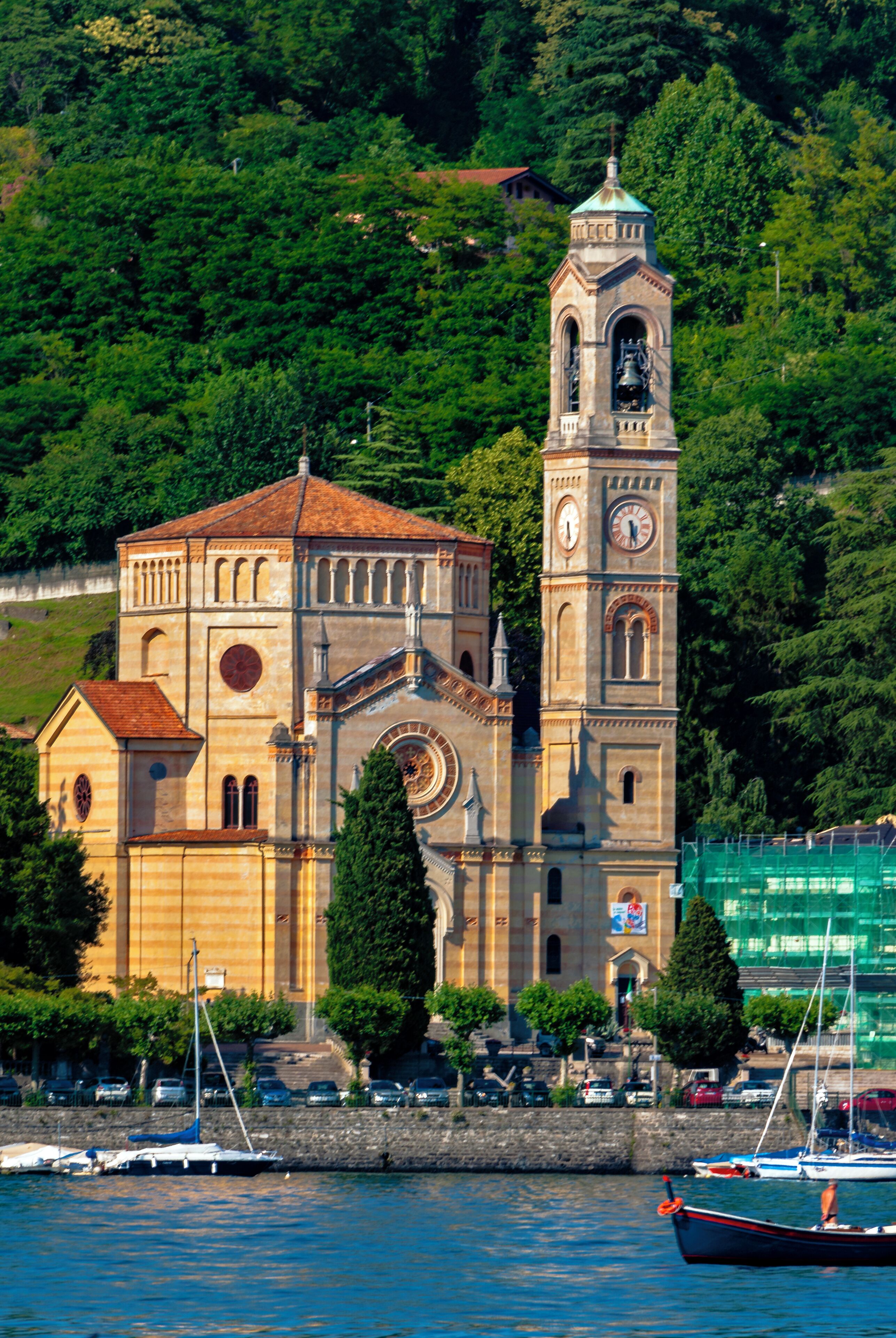 A historic church with a bell tower and a clock, surrounded by lush greenery and a body of water with boats.