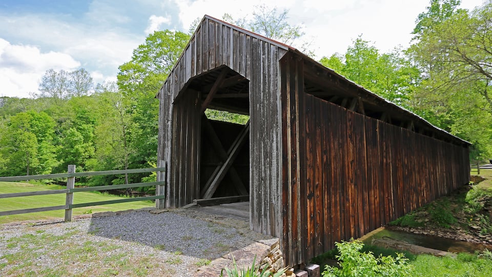 A wooden covered bridge with a stone foundation, surrounded by lush greenery and a wooden fence.