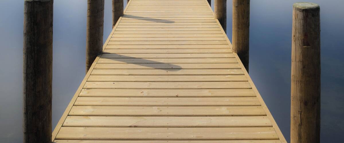A wooden pier extending into calm water, with a sailboat in the distance and hills in the background.