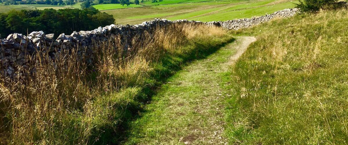 A stone wall, a grassy path, and a valley with fields and distant hills.