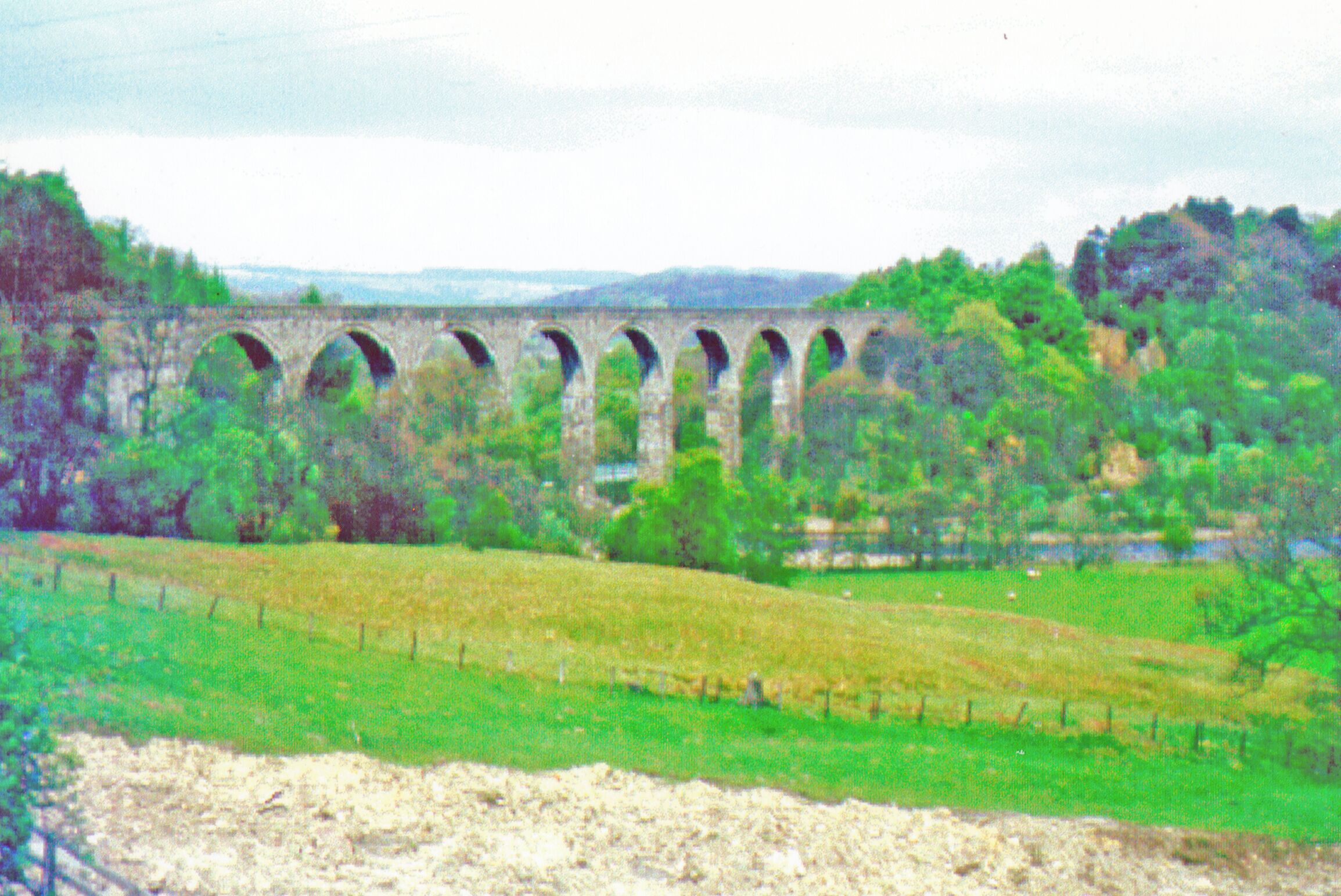 A stone viaduct with multiple arches over a river, surrounded by green fields and trees.