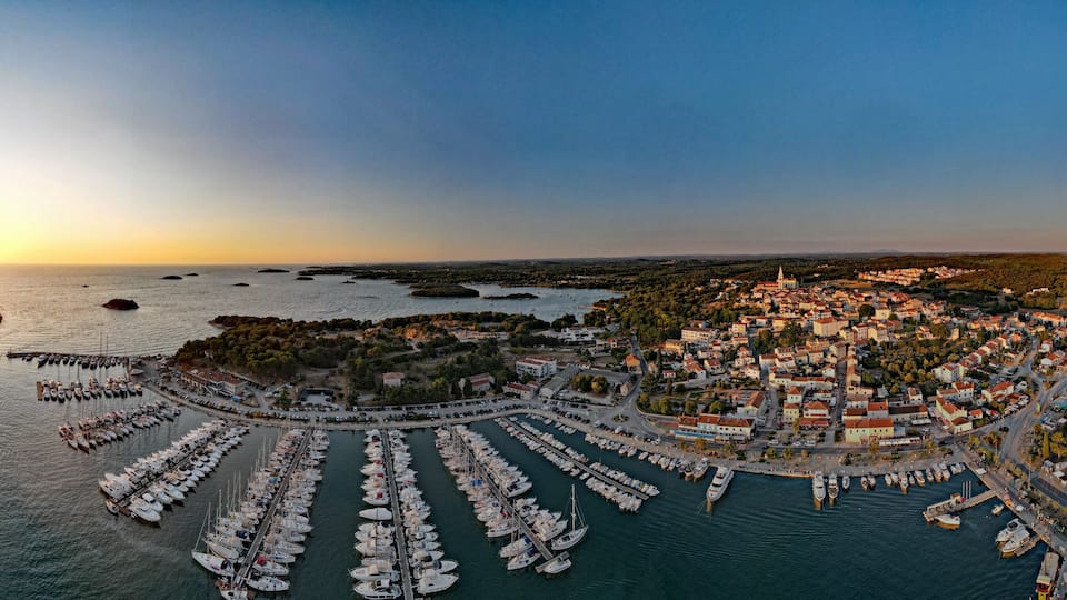 A coastal town with a marina filled with boats, a church spire, and a hilly landscape in the background.