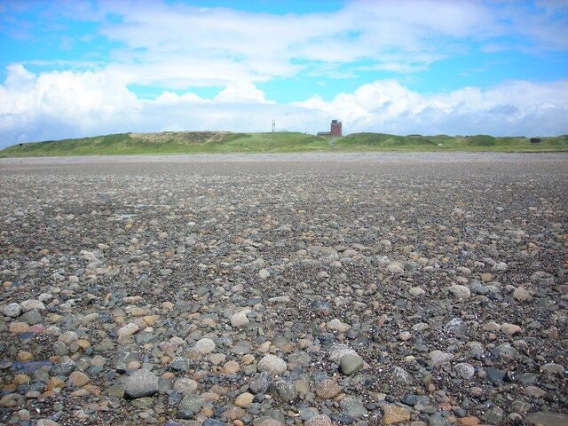 Uma praia de pedrinhas com um edifício distante numa colina gramada.