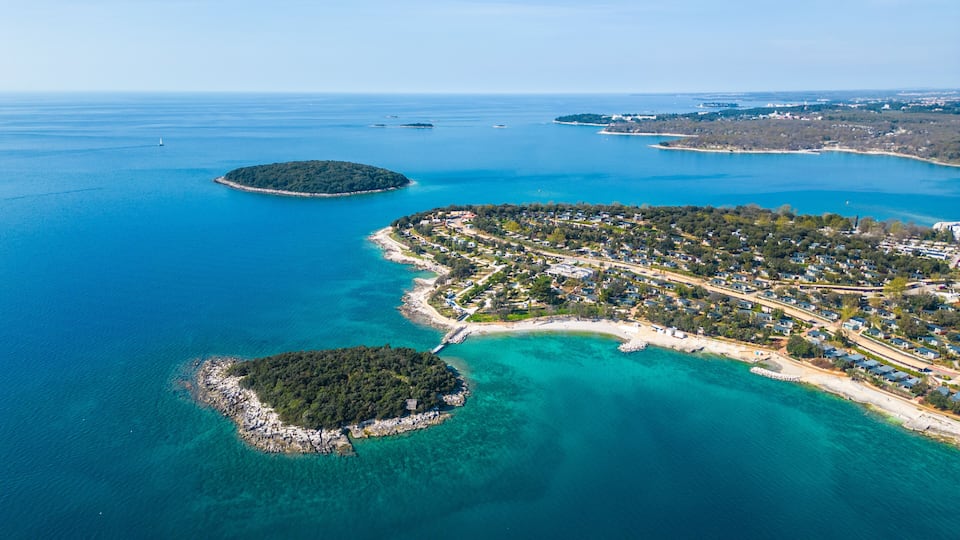 An aerial view of a coastal area with clear turquoise waters, a small island with dense vegetation, and a developed peninsula with residential buildings.