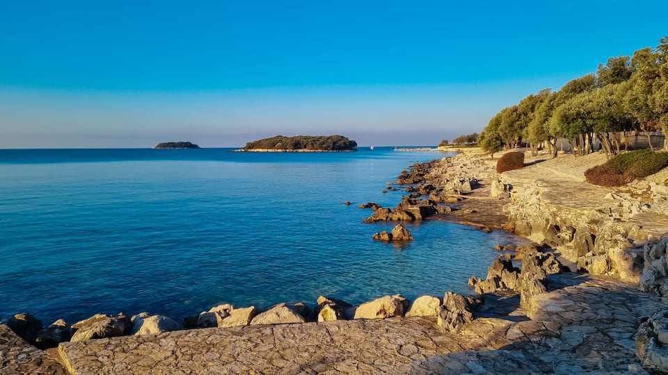 A coastal landscape with a rocky shoreline, a clear blue sea, and a small island in the distance.