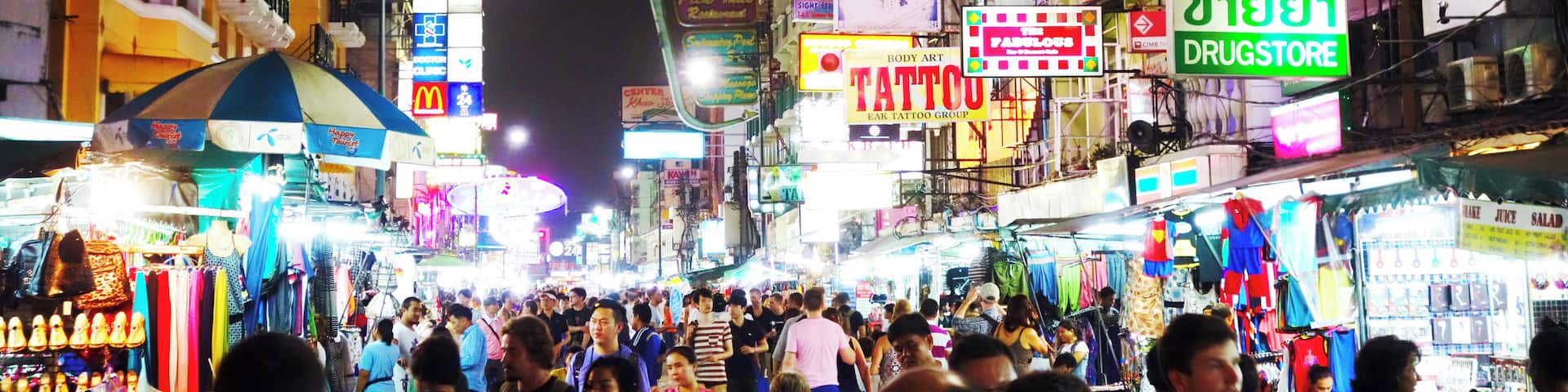 A crowded street at night with illuminated signs for a 7-Eleven, a tattoo parlor, and a drugstore.