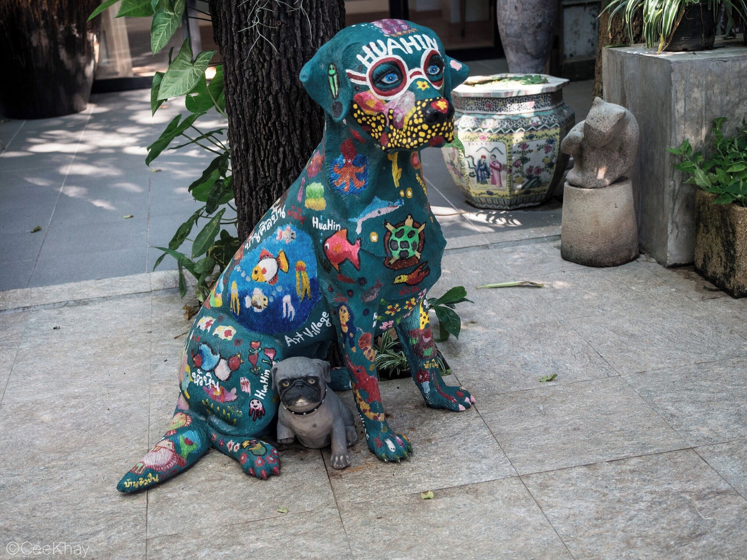 A colorful dog sculpture with a small dog beside it, set on a tiled surface with plants and a tree in the background.