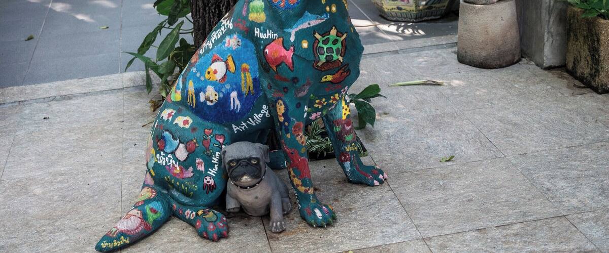 A colorful dog sculpture with a small dog beside it, set on a tiled surface with plants and a tree in the background.