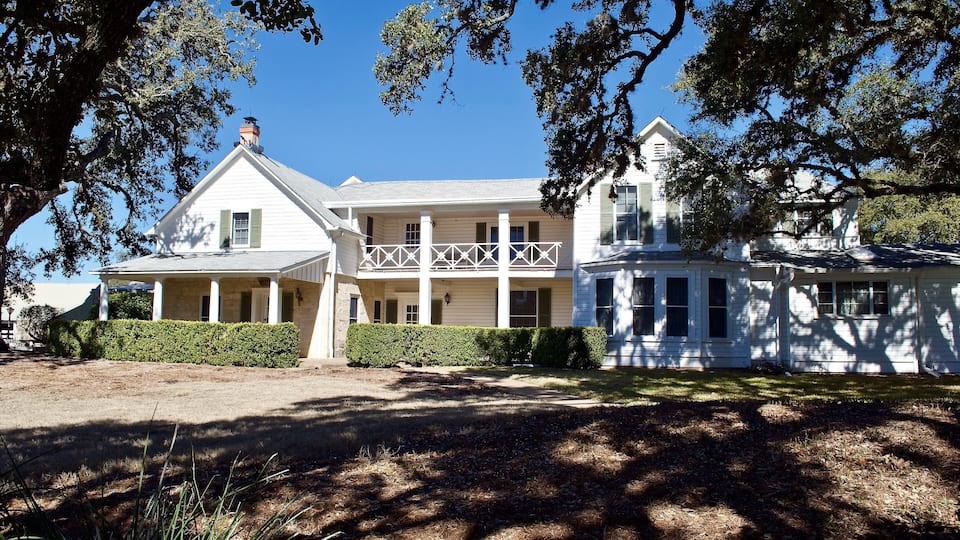 A large white house with a balcony, surrounded by trees and a well-maintained lawn.