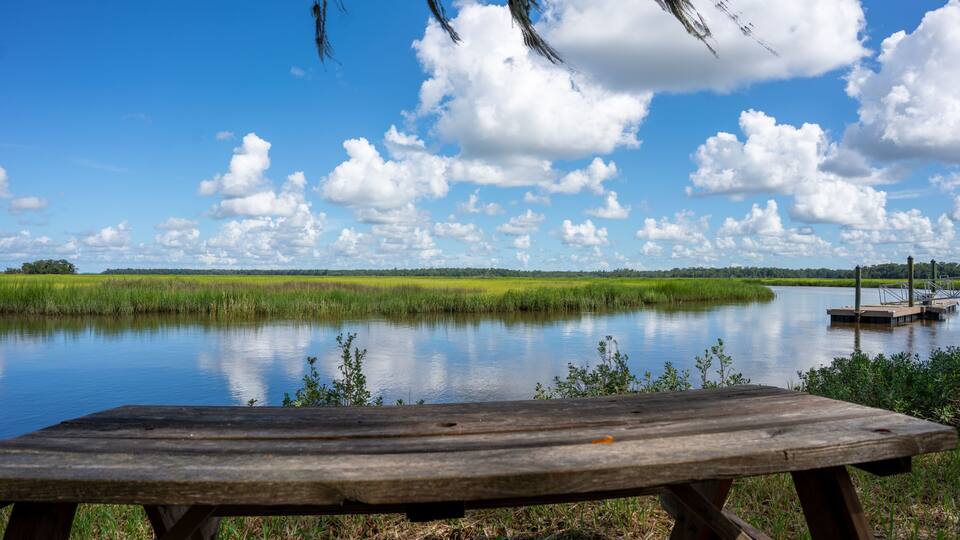A wooden picnic table by a calm lake with grassy banks and a dock in the distance.
