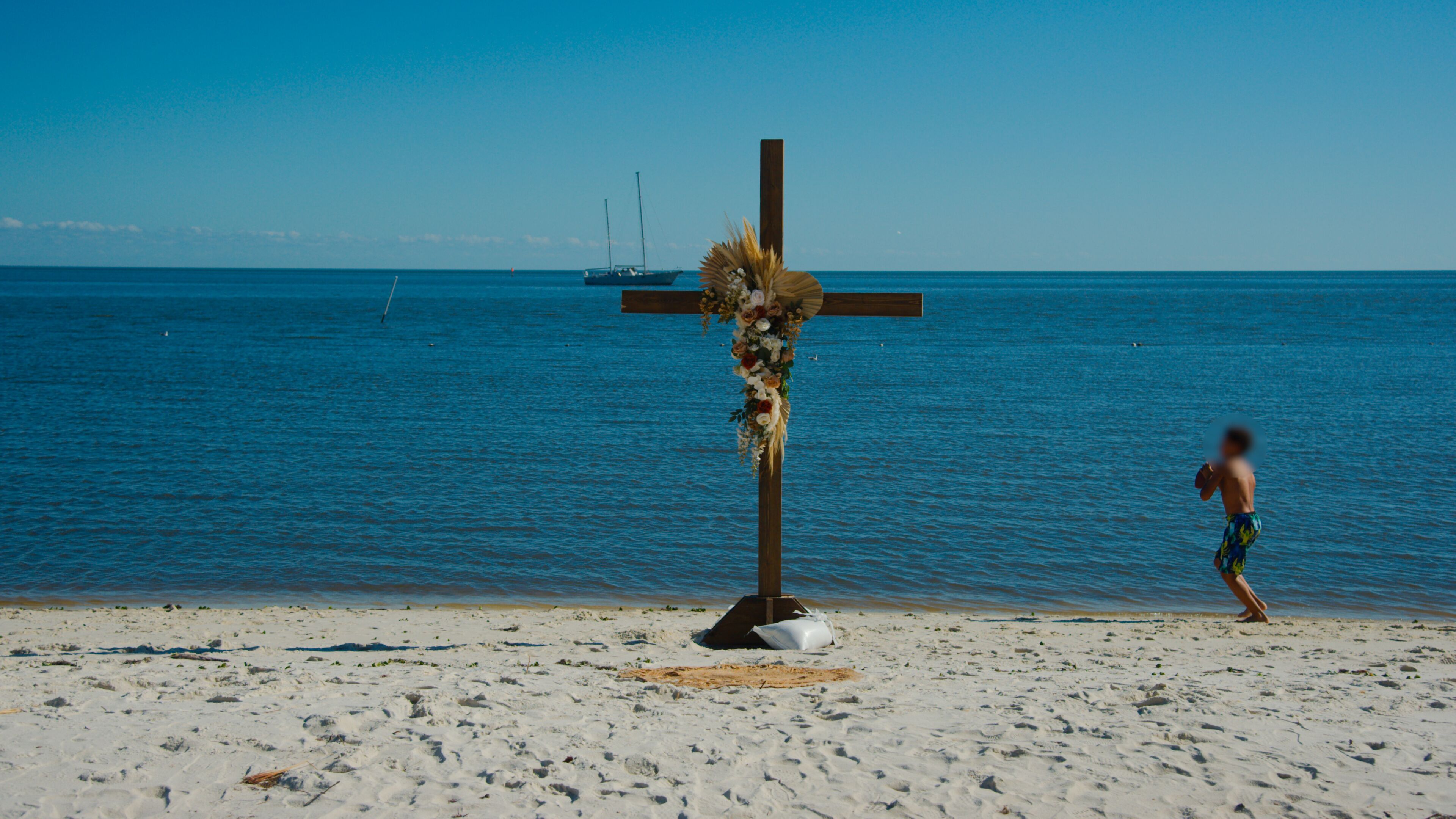 A beach with a cross adorned with flowers and a person walking.