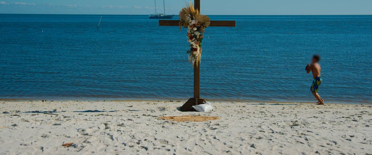 A beach with a cross adorned with flowers and a person walking.