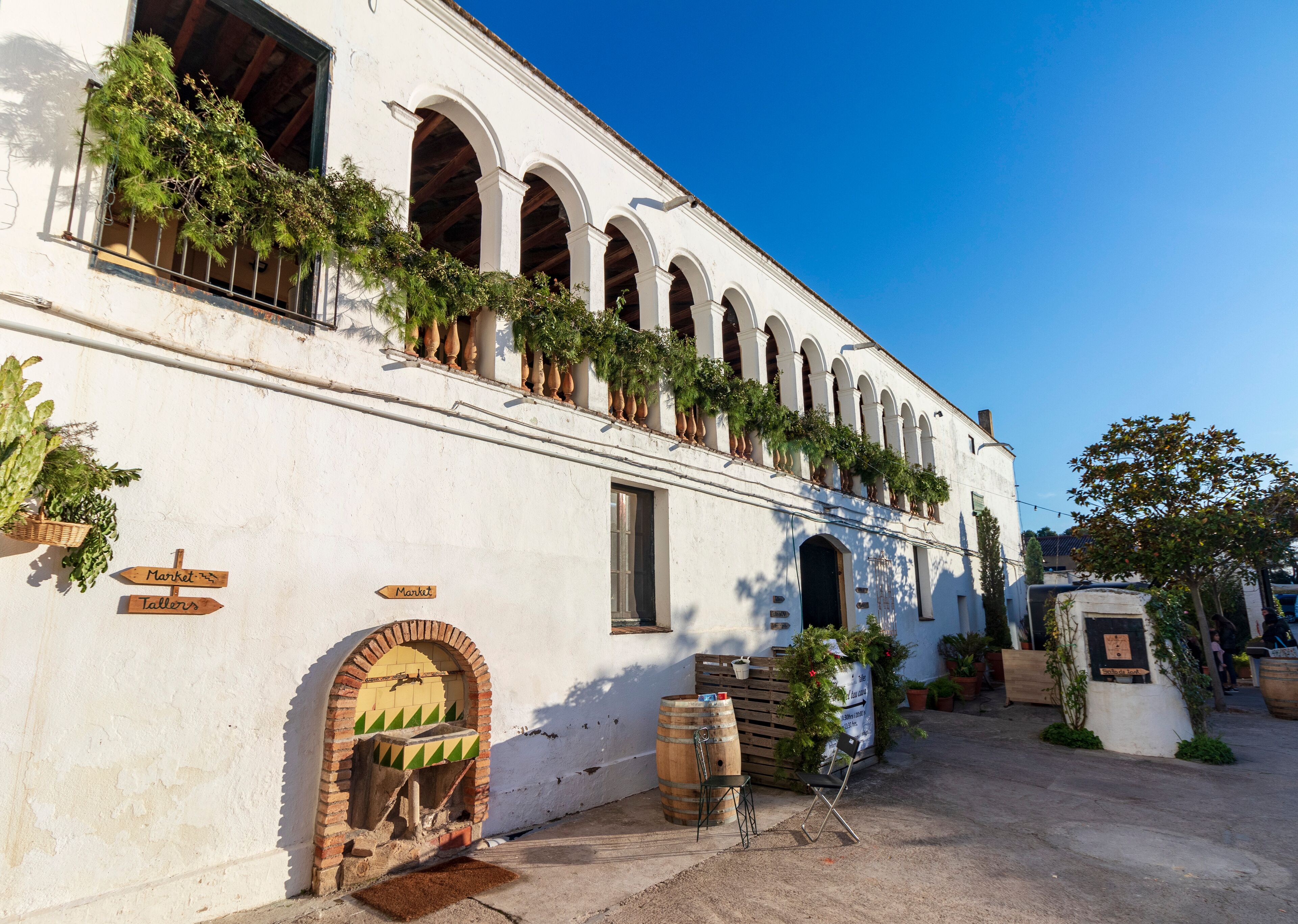 A white building with arched windows and a brick oven, surrounded by potted plants and outdoor seating.