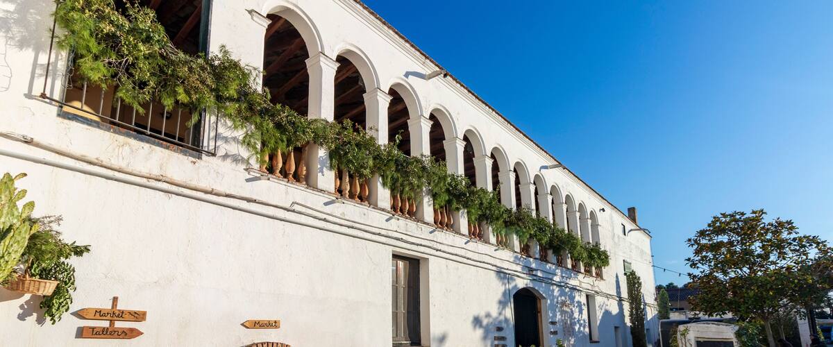 A white building with arched windows and a brick oven, surrounded by potted plants and outdoor seating.