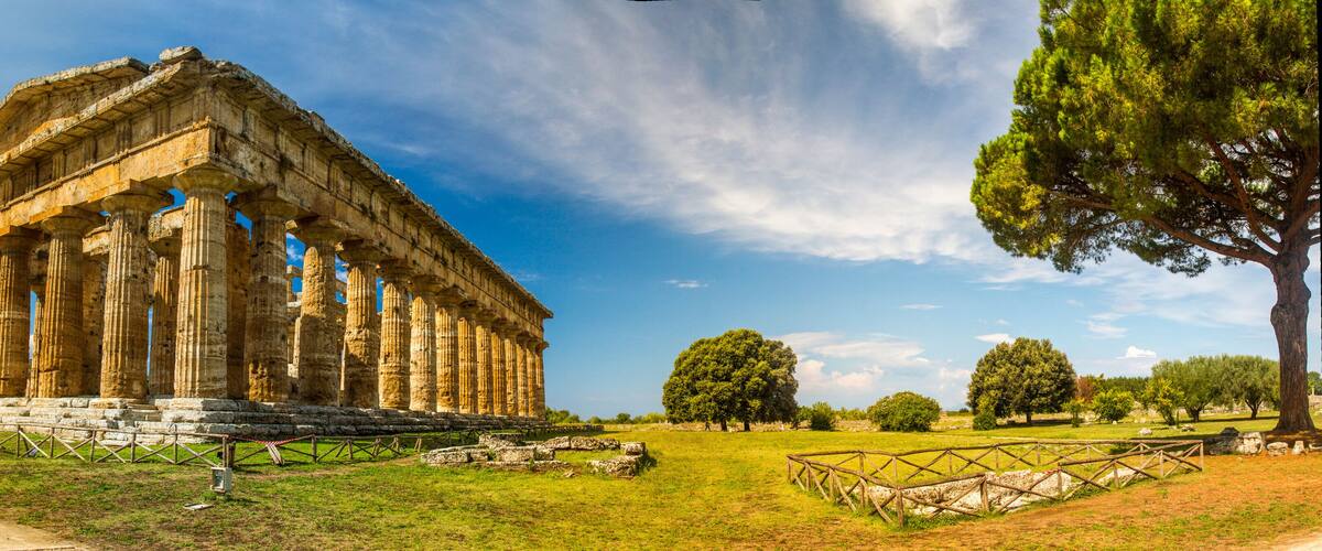 An ancient Greek temple with columns, surrounded by a grassy field and trees.