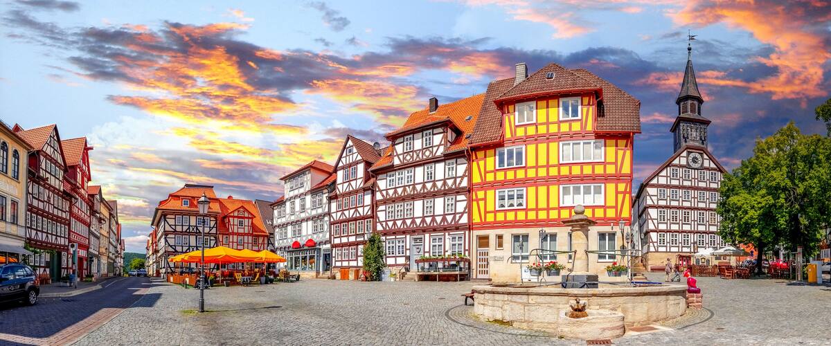 A cobblestone square with traditional half-timbered buildings, a clock tower, and a fountain.