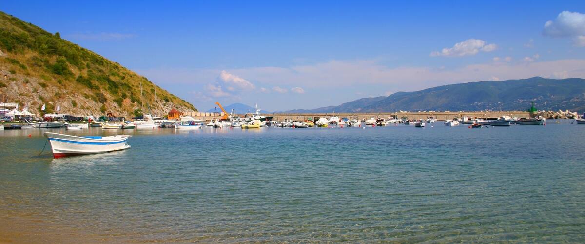 A coastal marina with boats docked, a sandy beach, and a hilly landscape in the background.