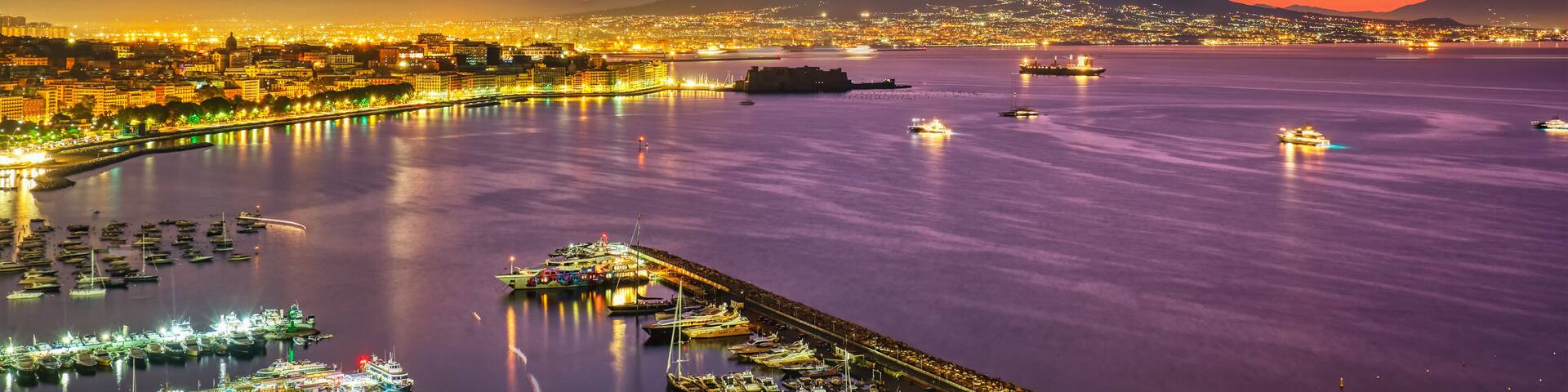 A coastal cityscape at dusk with illuminated buildings, a marina, and a mountain in the background.