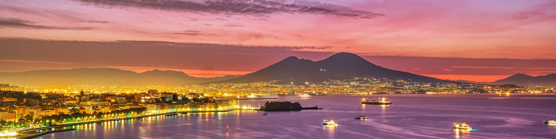 Un paisaje urbano costero al anochecer, con edificios iluminados, un puerto deportivo y una montaña en el fondo.
