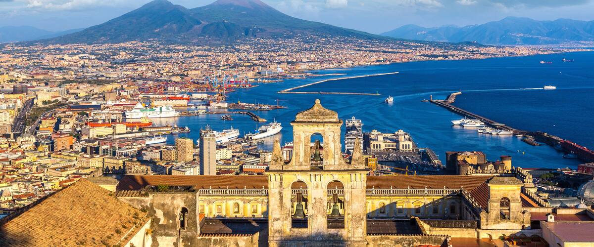 A coastal cityscape with a prominent cathedral, a large mountain in the background, and a busy harbor with ships.
