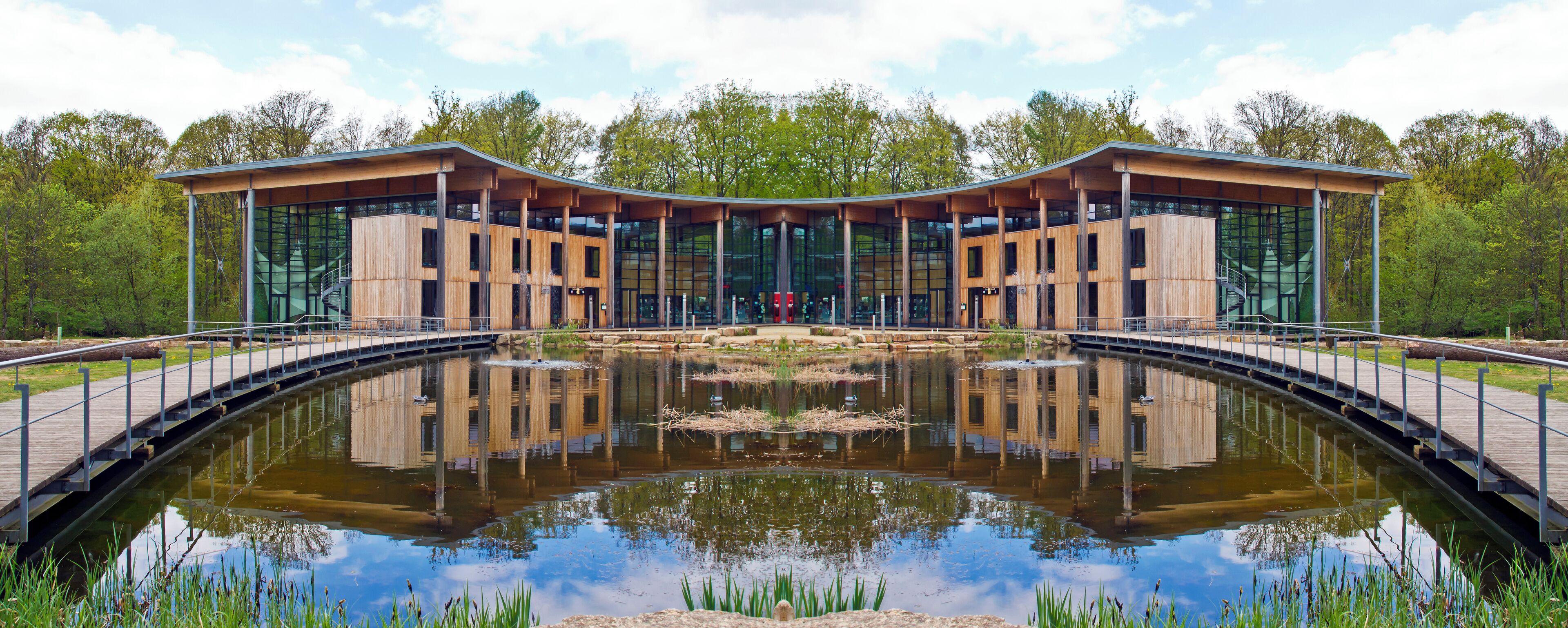 A modern building with a glass facade and wooden structure, surrounded by a reflective pond and greenery.