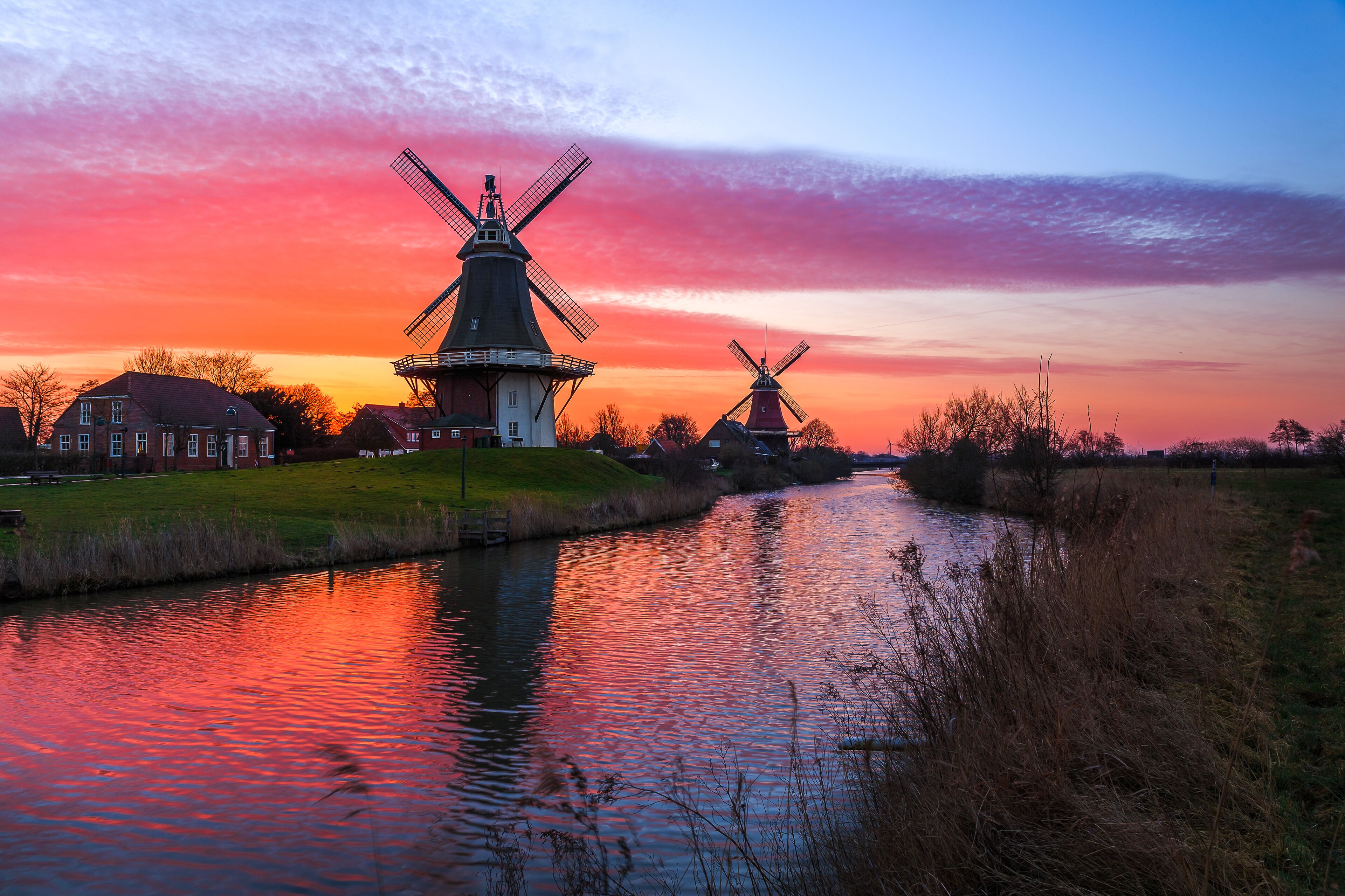 A canal with two windmills and a house in the background during sunset.