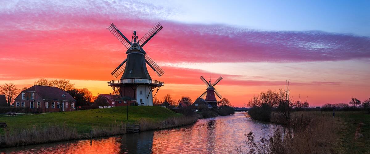 A canal with two windmills and a house in the background during sunset.
