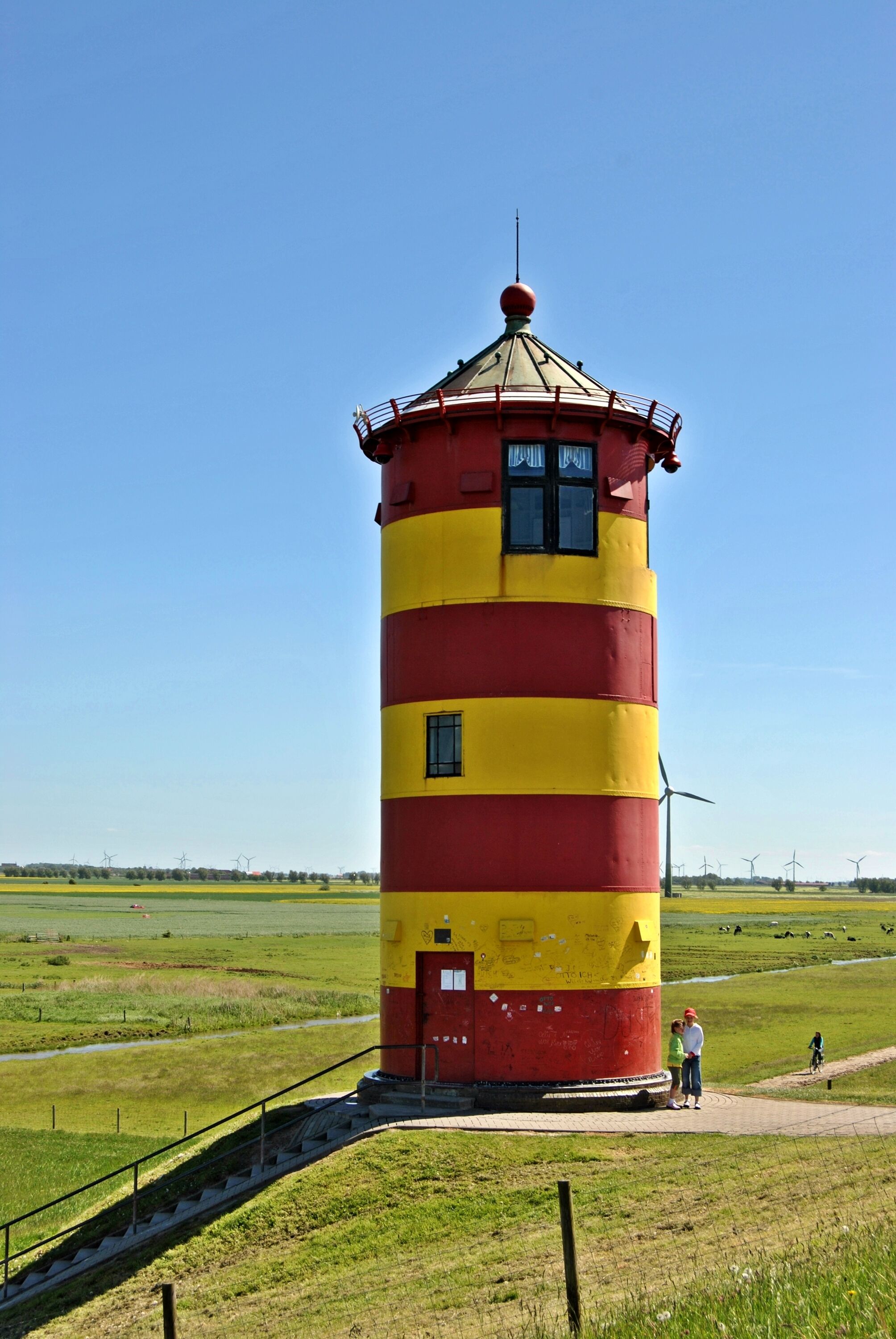 A tall, cylindrical lighthouse with alternating red and yellow stripes, situated in a grassy field with a clear sky.