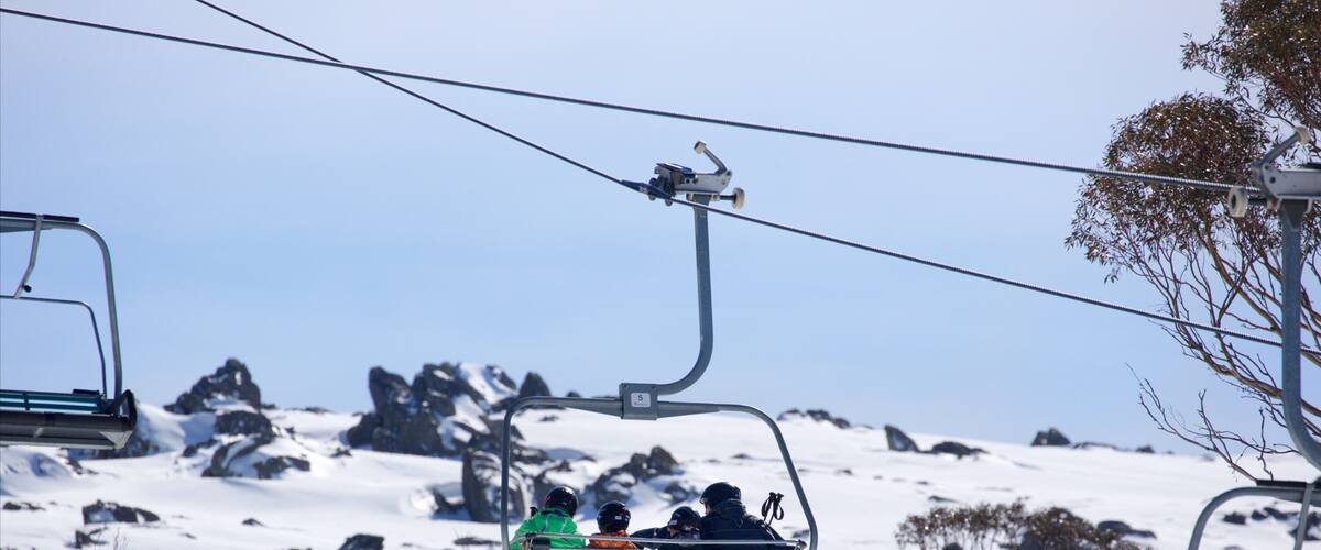 A ski lift with passengers, snow-covered mountains, and rocky terrain in the background.