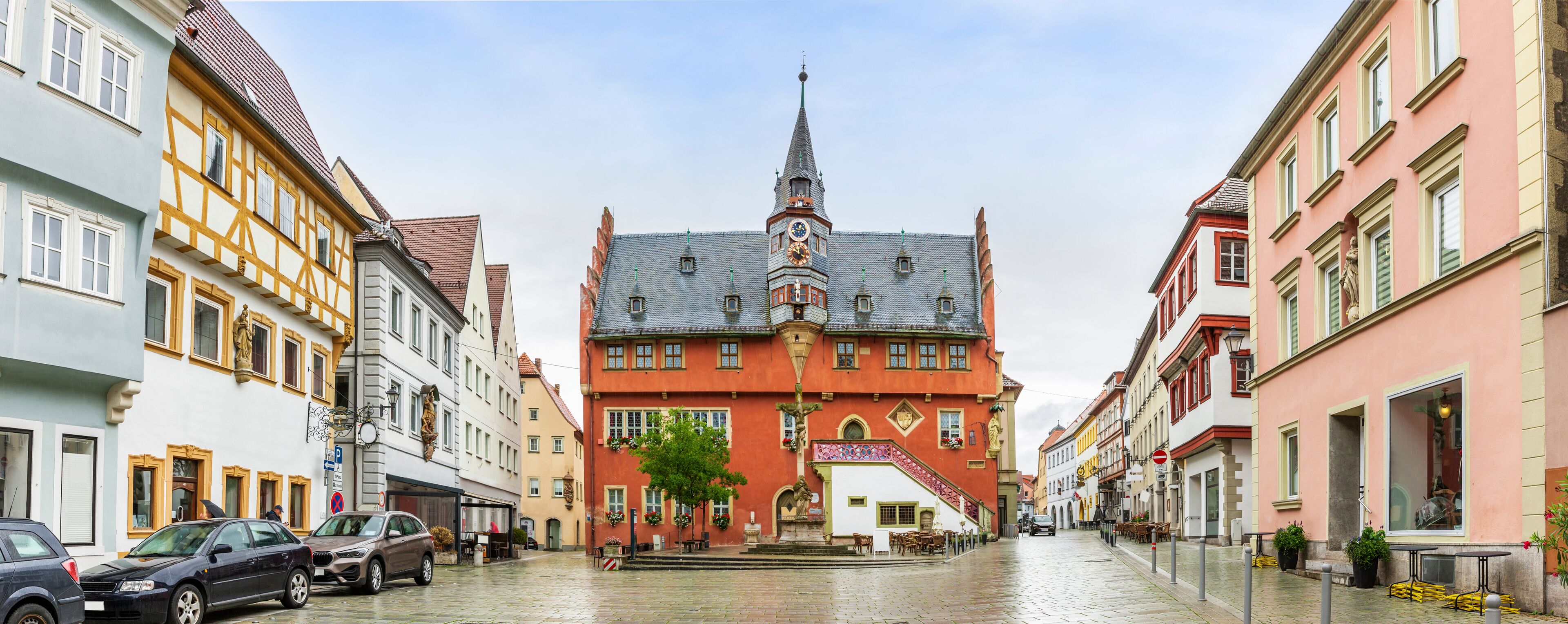A cobblestone street lined with historic buildings, a prominent church tower, and a small plaza with outdoor seating.