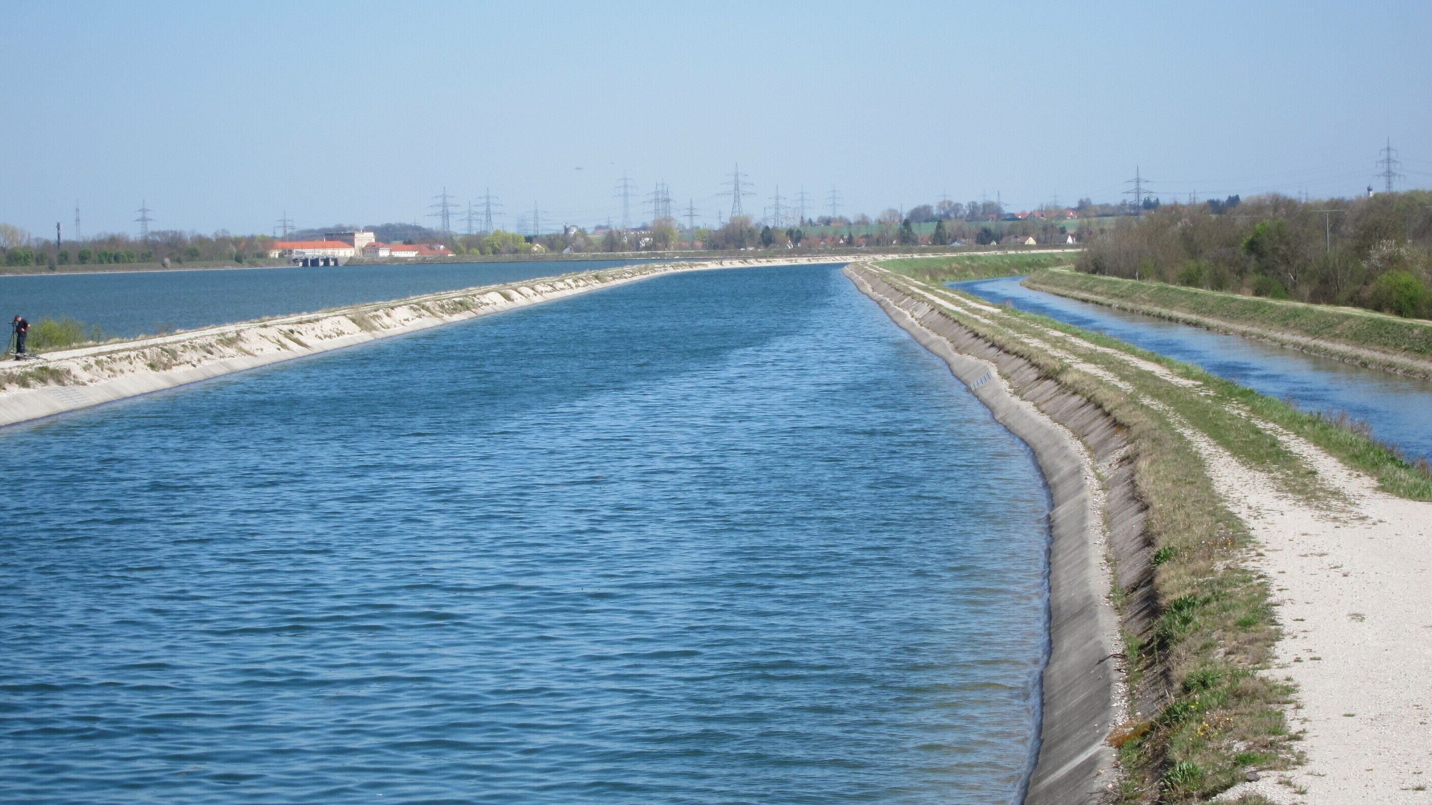 Der Mittlere-Isar-Kanal in der Mitte, links der Speichersee, rechts der deutlich tiefer liegende Entwässerungsgraben der Fischteiche. Alle drei Gewässer enden am Kraftwerk Neufinsing im Hintergrund.
