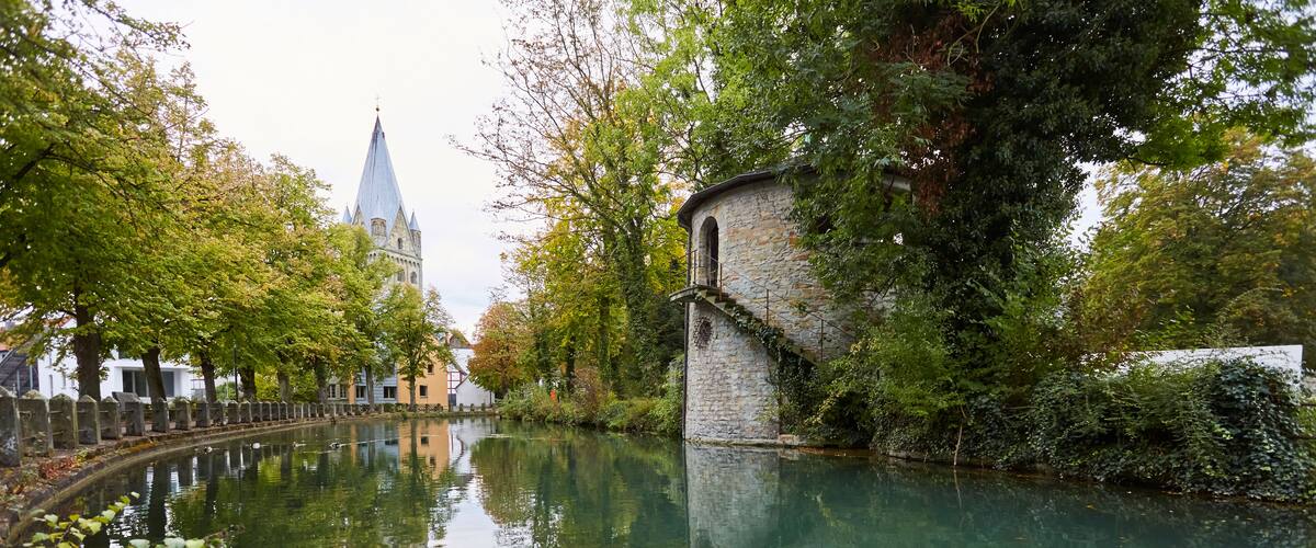 A calm river reflecting trees and a historic building with a tower.