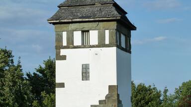 A stone tower with a white facade and a pointed roof, surrounded by a white picket fence and lush greenery.