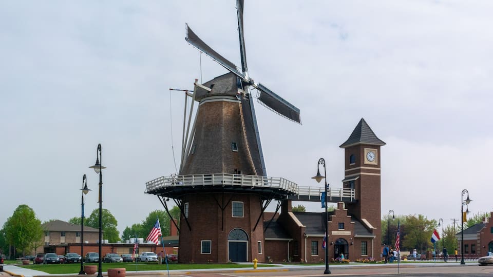 A historic windmill with a clock tower, surrounded by a park with American flags and a paved walkway.